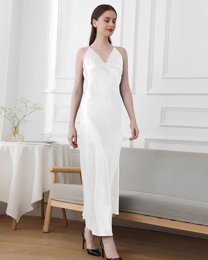 A woman stands in a white room wearing a Lace Long Silk Nightdress by SusanSilk. She is standing near a light-colored couch and table with a vase of flowers.