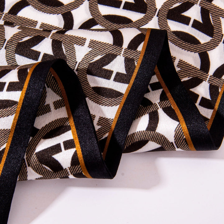 Close-up of the Ohm Pattern Narrow Long Silk Scarf by SusanSilk, showcasing a geometric pattern in black, white, and brown with a black and brown striped border along the edge. Crafted from luxurious silk, this intricate design brings both elegance and style to any ensemble.