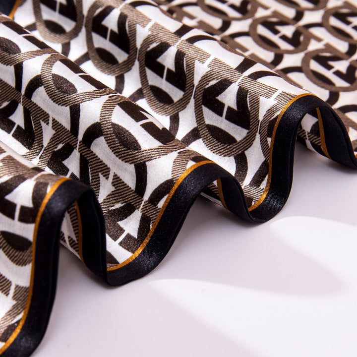 The Ohm Pattern Narrow Long Silk Scarf from SusanSilk, with its black and brown geometric pattern featuring interlocking shapes, is draped in undulating waves on a white surface, showcasing its elegance.