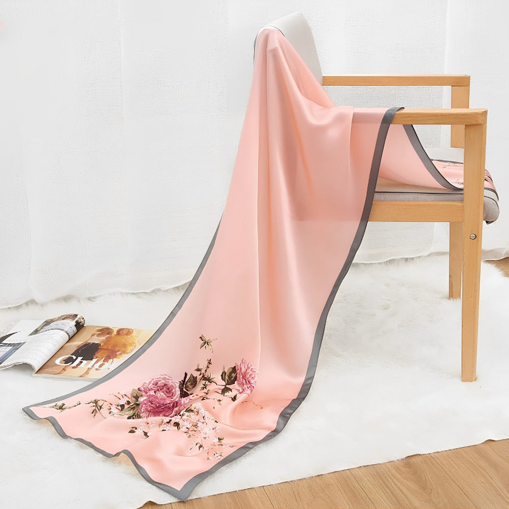 An Ornament & Floral Long Silk Crepe Scarf by SusanSilk, featuring pink floral patterns with a gray border, draped over a wooden chair on a white rug, next to an open magazine.