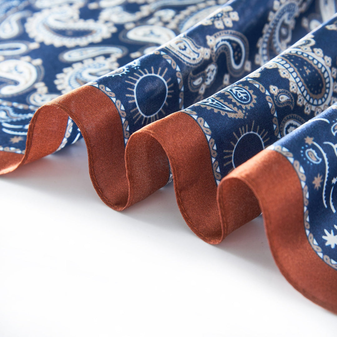 A close-up of the luxurious Paisley Series Square Silk Scarf by SusanSilk, showcasing its intricate blue, white, and brown paisley pattern. The silky material is gracefully folded in a wave-like pattern.