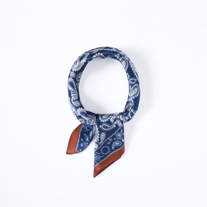 A SusanSilk Paisley Series Square Silk Scarf, featuring a blue paisley pattern accented with brown edges, neatly rolled into a circular shape against a white background.