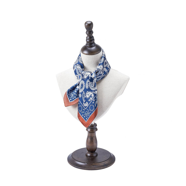 A mannequin showcases the Paisley Series Square Silk Scarf by SusanSilk, featuring a blue paisley pattern with brown edges, crafted from luxurious silk and elegantly draped and tied around its neck.