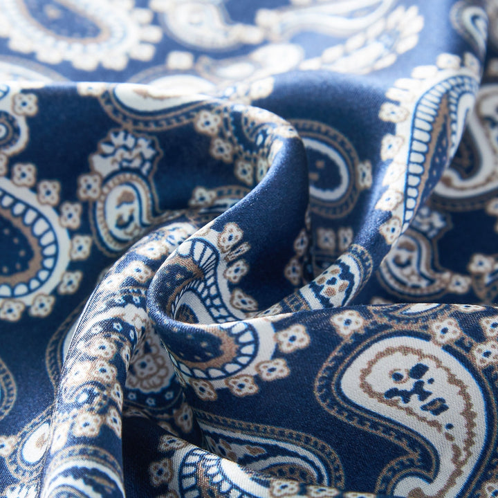 Zoomed-in view of a SusanSilk Paisley Series Square Silk Scarf showcasing a blue background adorned with intricate white and light brown paisley patterns. The silk fabric looks soft and delicately folded.