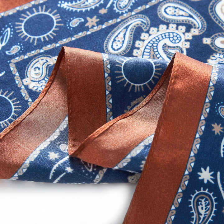 Close-up of a fabric from the SusanSilk Paisley Series Square Silk Scarf, showcasing a blue and white paisley design adorned with suns and stars, and bordered with a brown satin ribbon. This elegant piece is crafted from fine silk, adding an extra touch of luxury.