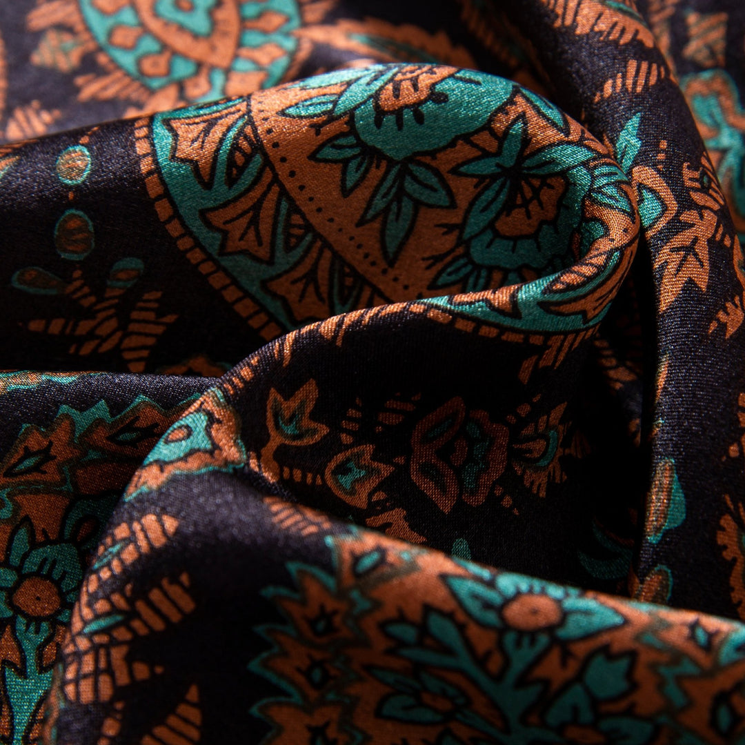 Close-up of the Paisley Square Silk Scarf - Retro by SusanSilk, showcasing an intricate floral pattern in shades of teal and orange on a black background. The slightly crumpled silk fabric highlights its luxurious texture.