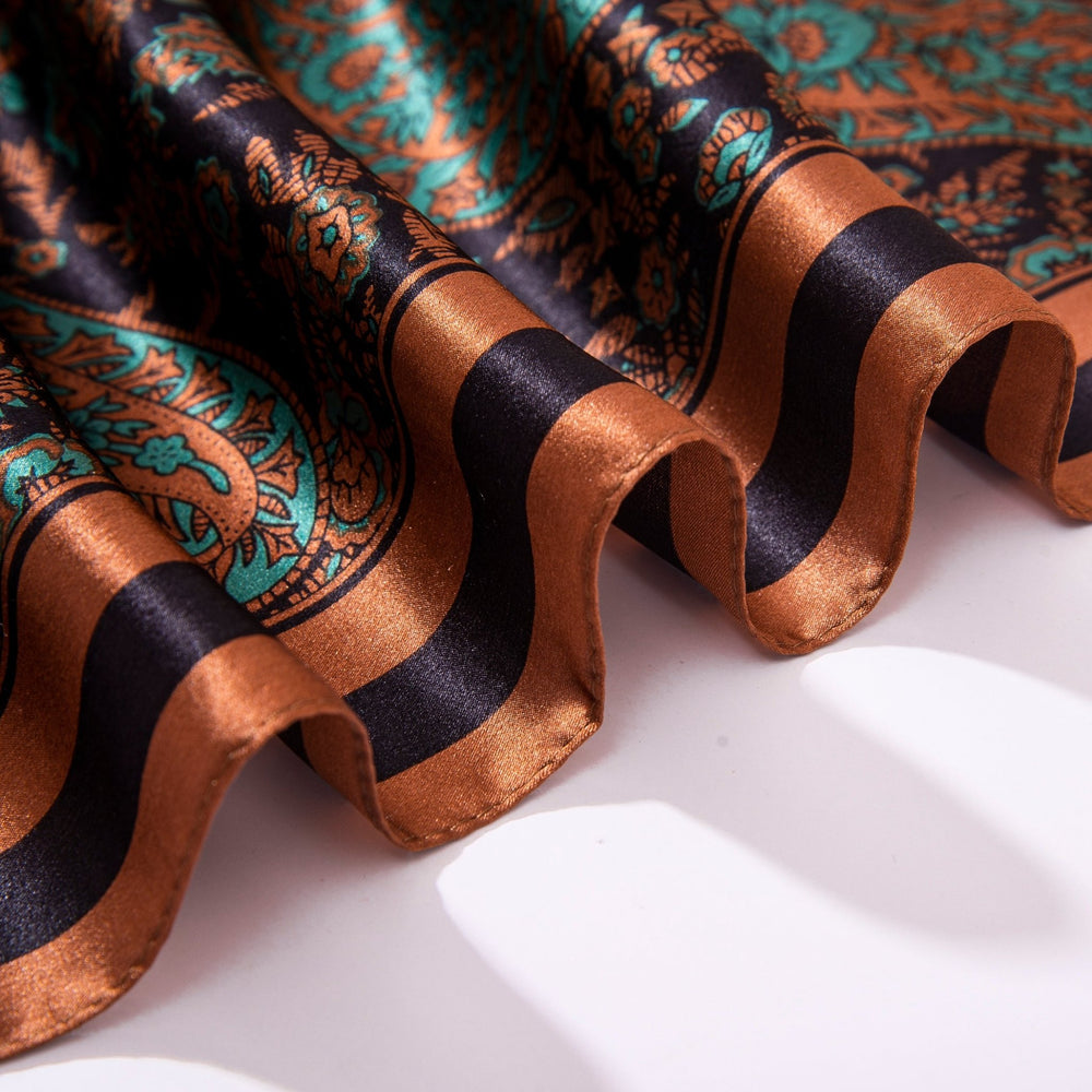 Close-up of a folded Paisley Square Silk Scarf - Retro from SusanSilk, showcasing a detailed, intricate pattern in shades of copper, black, and teal. The scarf's glossy texture and design are beautifully illuminated under soft lighting.